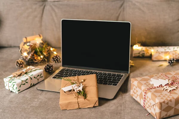 Online αγορές στις διακοπές των Χριστουγέννων. Δώρα, και mock up οθόνη laptop στον καναπέ με φυσικά οικολογικά δώρα και διακόσμηση. Καλά Χριστούγεννα. Περιτύλιγμα των παρόντων κιβωτίων. — Φωτογραφία Αρχείου