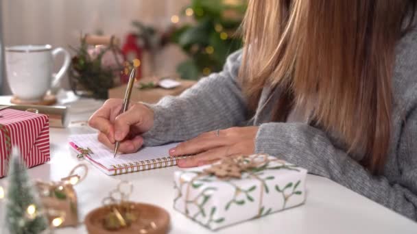 Hedefler, yılbaşı konsepti için kız defterine yazma ve dilek listesi hazırlar. Kış tatilinde evde elinde kalemle gezen bir kadın. Noel süsü, hediye kutuları. — Stok video