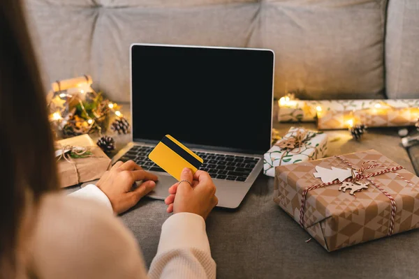 Закріпіть руки з кредитною карткою, подарунками і висмійте екран ноутбука. Інтернет-магазин на Різдвяних канікулах. Розпечена самиця сидить на дивані з природними подарунками еко та декором. Веселого Різдва — стокове фото