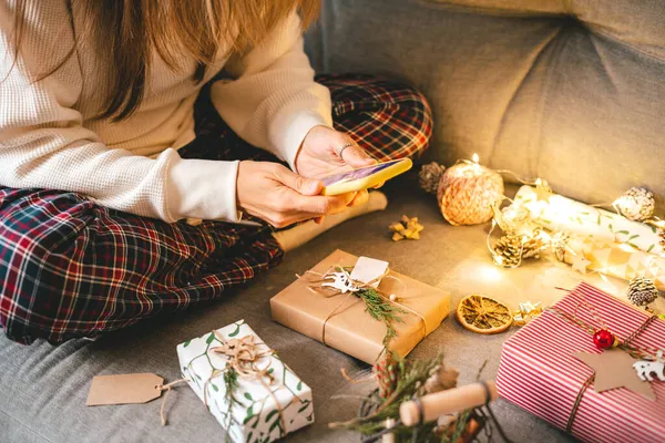 Mujer con teléfono envolviendo cajas de regalo de Navidad, tomando fotos. Mujer recortada sentarse y preparar regalos ecológicos naturales en el sofá con elementos de decoración. Feliz Navidad o Año Nuevo DIY embalaje Concepto — Foto de Stock