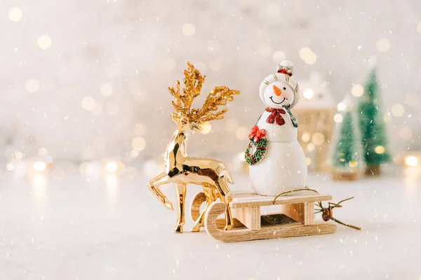 Golden deer dragging snowman on toy wooden sled on gray background with glowing light bokeh, copy space. New year card. Xmas decoration with Christmas tree, house and snow — Stock Photo, Image