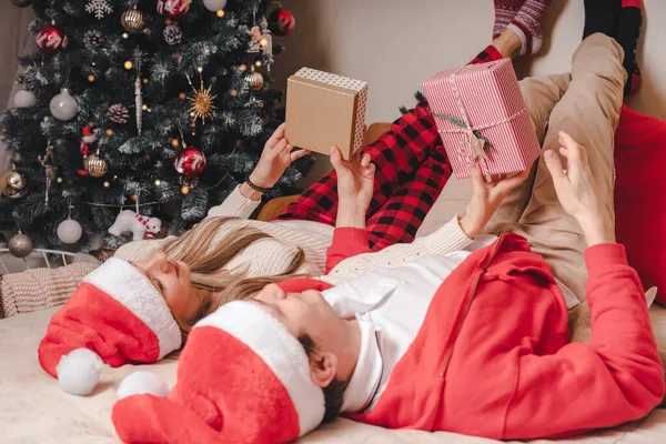 Ρομαντικό ζευγάρι ανταλλαγή δώρων κοντά διακοσμημένο χριστουγεννιάτικο δέντρο. Άνδρας που δίνει χριστουγεννιάτικο δώρο έκπληξη σε χαμογελαστή γυναίκα με πουλόβερ ενώ χαλαρώνει στον καναπέ. Φίλος και φίλη. — Φωτογραφία Αρχείου