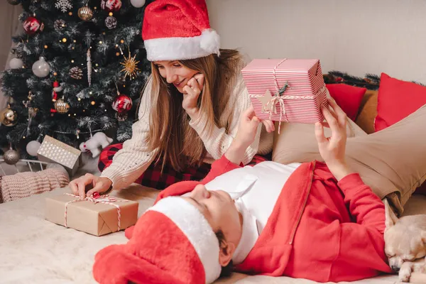 Pareja romántica intercambiando regalos cerca del árbol de navidad decorado. Hombre dando regalo de Navidad sorpresa a la mujer sonriente en suéter mientras se relaja en el sofá. Novio y novia. — Foto de Stock