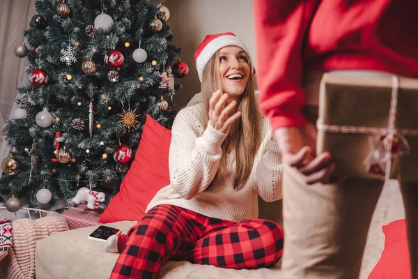 Sorprendente... Vista trasera de cerca del hombre en suéter rojo sosteniendo una caja de regalo de Navidad a sus espaldas mientras está de pie cerca del árbol de Navidad decorado. Pareja familiar en vacaciones de Navidad. — Foto de Stock
