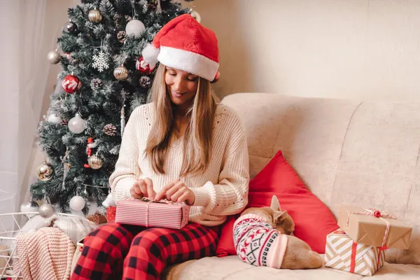 Γυναίκα σε Σάντα καπέλο περιτύλιγμα κουτιά δώρο Χριστουγέννων με κουτάβι σκυλί σε πουλόβερ. Γυναίκα κάθονται και την προετοιμασία δώρα στον καναπέ με διακόσμηση και χριστουγεννιάτικο δέντρο. Καλά Χριστούγεννα ή το Νέο Έτος DIY συσκευασίας Concept — Φωτογραφία Αρχείου