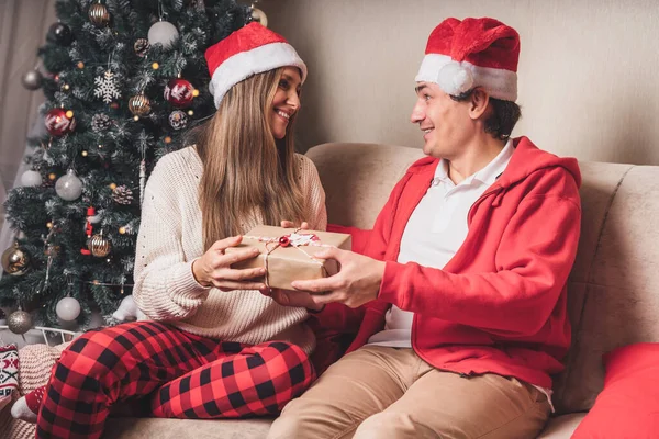 Romantic couple exchanging gifts near decorated christmas tree. Man giving christmas present surprise to smiling woman in sweater while relaxing on couch. Boyfriend and girlfriend. — Stock Photo, Image
