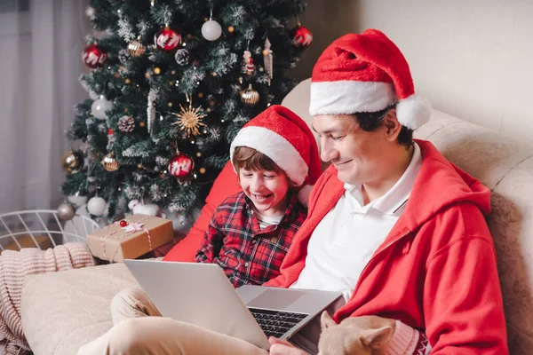 Padre con hijo niño hijo y cachorro perro en el sombrero de Santa en las vacaciones de Navidad, sentado en un sofá, tener un video chat en el ordenador portátil en la sala de estar con árbol de Navidad en casa. Chicos ver película. — Foto de Stock