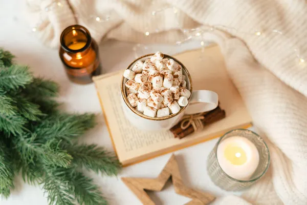 Hygge fincan sıcak kakao ya da şekerli kahve, kazak, mum, kitap, tarçın ve köknar ağacı. Noel ışıkları rahat arka plan. Noel tatili ahşap yıldız süslemeleri