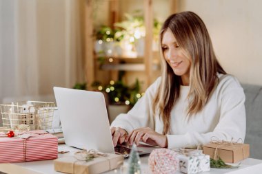 Mutlu ve gülümseyen bir kadın Noel hediyelerini paketler ve hazırlar, dizüstü bilgisayarda yazar ve görüntülü konuşma yapar. Noel tatilinde online alışveriş. Defterle evden uyanan serbest kız.