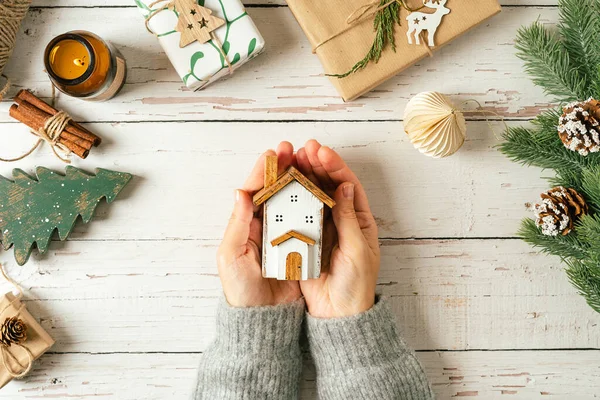Flat lay of woman hands holding cozy homemade wooden house on white background with Christmas natural eco decoration. Top view gifts, candle, fir tree, toy, cinnamon, pine cone, craft