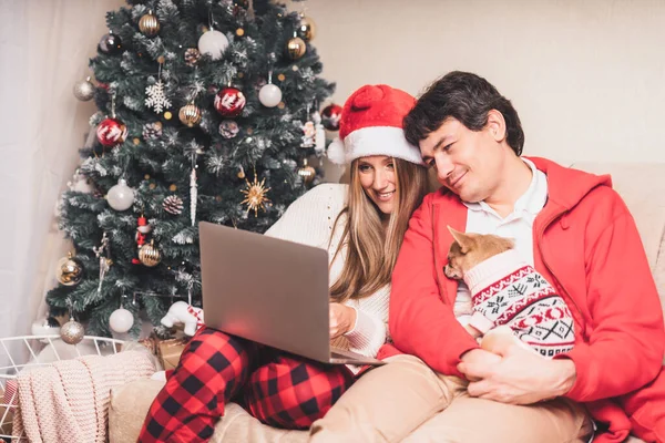 Πορτρέτο της υπέροχο ζευγάρι σύζυγος και η σύζυγος με φορητό υπολογιστή σε κόκκινο καπέλο Σάντα έχοντας ένα βίντεο κλήση chat με κουτάβι σκυλί σε πουλόβερ, να απολαύσετε τα Χριστούγεννα στο σπίτι. Online χαιρετισμός. — Φωτογραφία Αρχείου