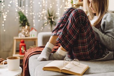 Sıcak çorap, kazak ve kareli ekose çorap giymiş sıcak kakao ya da kupada kahve içen evde kanepede dinlenirken kitap okuyan rahat kadın bacakları. Dekor ve ışıklarla Noel tatili.
