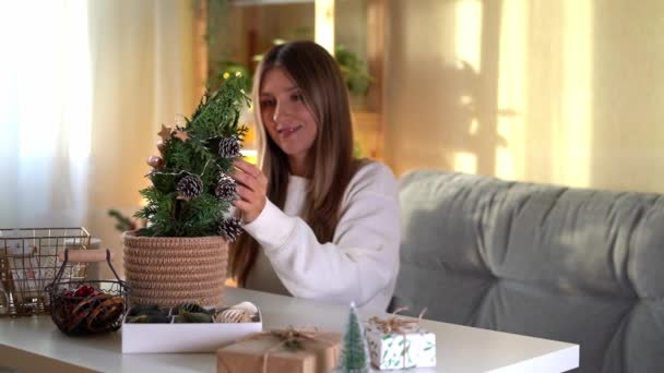 Nő díszítő öko természetes karácsonyfa pot otthon. Ajándék dobozok elkészítése fehér asztalon díszítő elemekkel, fényekkel és tárgyakkal Karácsonyi vagy újévi barkácsolás koncepció. — Stock videók