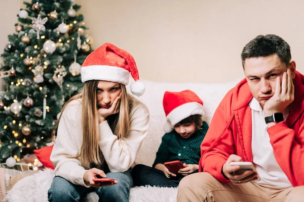 Portret przygnębionej rodziny z synem bawiącym się komórką podczas imprezy noworocznej. Zmęczeni mama i tata spędzają stresujący czas podczas gdy dziecko używa telefonu komórkowego. Spór rodzinny po świętach Bożego Narodzenia — Zdjęcie stockowe