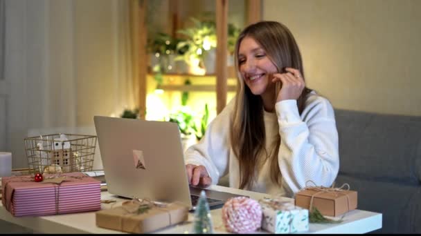 Gelukkige en lachende jonge vrouw inpakken en voorbereiden van kerstcadeaus, met behulp van laptop en het hebben van online video chat. Online winkelen op kerstvakantie. Huis interieur met verlichting en planten. — Stockvideo