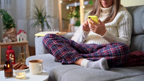 Άνετο γυναίκα σε πλεκτό ζεστό κάλτσες χειμώνα, πουλόβερ και καρό καρό καρό με τηλέφωνο, πίνοντας ζεστό κακάο ή καφέ σε κούπα, κατά τη διάρκεια ανάπαυσης στον καναπέ στο σπίτι. Χριστουγεννιάτικες διακοπές με κερί, διακόσμηση και μπισκότο — Αρχείο Βίντεο