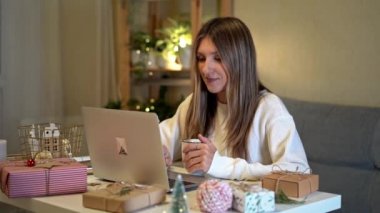 Mutlu ve gülümseyen genç bir kadın Noel hediyelerini paketliyor ve hazırlıyor, dizüstü bilgisayar kullanıyor ve çevrimiçi görüntülü sohbet ediyor. Noel tatilinde online alışveriş. Işıklar ve bitkilerle dolu bir ev..