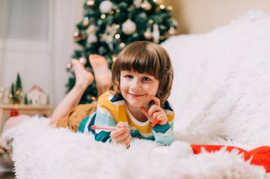 Gülümseyen çocuk kanepeye uzanıp Noel ağacının yanındaki Noel Baba 'ya mektup yazıyor. Çocuk dilek listesi. Noel hediyelerinin rüyaları. Mutlu Noeller ve mutlu yıllar..