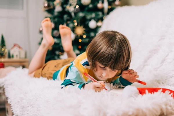 Χαμογελαστό παιδί ξαπλωμένο στον καναπέ και γράφοντας το γράμμα στον Άγιο Βασίλη στο σπίτι κοντά στο χριστουγεννιάτικο δέντρο. Λίστα επιθυμιών παιδιών. Όνειρα χριστουγεννιάτικων δώρων. Καλά Χριστούγεννα και Ευτυχισμένο το νέο έτος. — Φωτογραφία Αρχείου