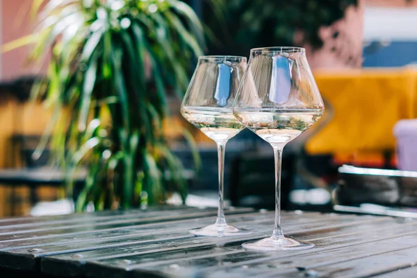 A wooden table in a restaurant with white wine. Wine glasses served for a party in a bar or a restaurant on terrace at a sunny day. Blurred background with plants. Still life scene. — Stock Photo, Image