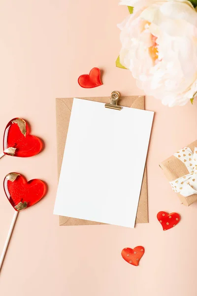 Colocación plana de sobre de papel con tarjeta de papel de felicitación en blanco. Fondo de mesa rosa con regalo de San Valentín, carta, forma de corazón, flores, piruletas y decoración. Vista superior, simulacro de invitaciones. — Foto de Stock