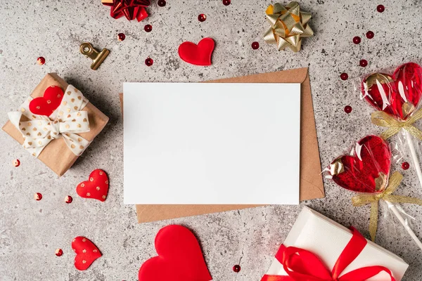 Colocación plana de sobre de papel con tarjeta de felicitación en blanco. Fondo de mesa gris con regalo de San Valentín, carta, forma de corazón, confeti, piruletas y decoración. Vista superior, simulacro de invitaciones. — Foto de Stock