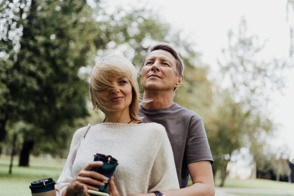 An adult mature happy couple in love hugging outdoors in city park. A blonde caucasian man and woman spend time together and drinking coffee. Senior wife and husband walking outside.