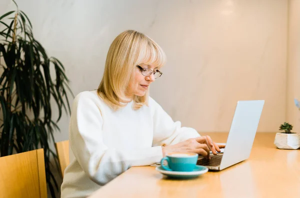 Yetişkin bir kadın kahve fincanıyla kafede oturuyor ve internette dizüstü bilgisayarla çalışıyor. Gözlüklü bir iş kadını, kafeteryadaki ortak çalışma alanında defter bilgisayarında daktilo kullanıyor. — Stok fotoğraf