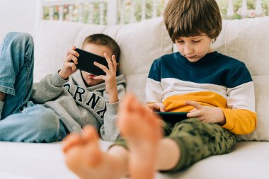 Neşeli iki çocuk online oyun oynuyor, cep telefonundan video izliyorlar. Komik mobil uygulamalar kullanarak gülümseyen çocuklar, kanepede boş vakitlerin tadını çıkarıyorlar. Kardeşler telefonda birlikte oynarlar.