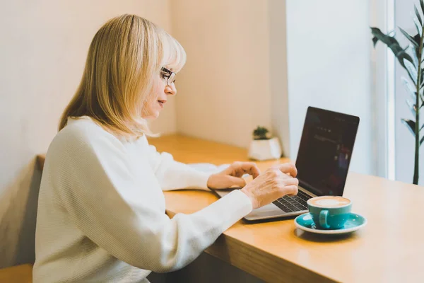 Gammal vuxen kvinna sitter i café med kaffemugg och arbetar online på bärbar dator. Affärskvinna i glasögon skriva på bärbar dator i coworking utrymme i rostar kafé — Stockfoto