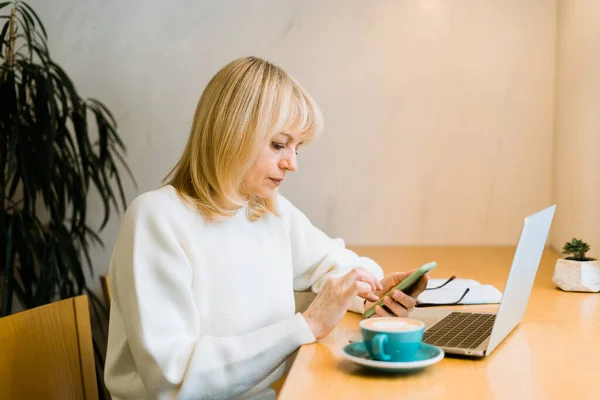 Yetişkin bir kadın kahve fincanıyla kafede oturuyor ve internette dizüstü bilgisayarla çalışıyor. İş kadını, taşlama kahve dükkanındaki ortak çalışma alanındaki cep telefonuna mesaj atıyor. — Stok fotoğraf