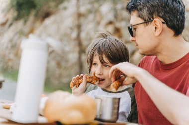 Babasının yakın çekim görüntüsü, dağlarda bir aile pikniğinde okul oğluna sarılıyor. Çocuk ve babası dağlarda yürüyüş yaparken dinlenip pikniğin tadını çıkarıyorlar.