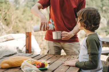 Father dad pours hot coffee tea from thermos into the mug on a family picnic in the mountains. Child school boy kid is watching his dad filling the cup with hot beverage