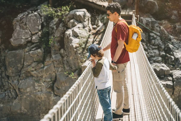 Vater und Sohn schauen beim Wandern von der Hängebrücke in die Berge. Lässig gekleidete Touristen, Schulkind und sein Vater mit gelbem Rucksack überqueren die Schlucht durch eine Hängebrücke — Stockfoto