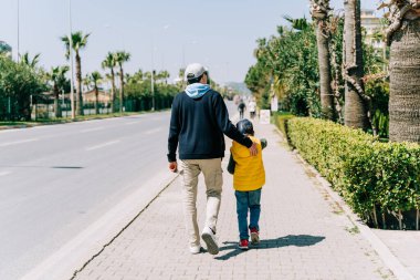 Rearview of father and son walking near city road. Dad holding his kids back when taking a stroll in the southern city. Man and boy going for promenade near highway