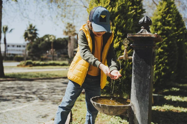 Kid schoolboy in casual clothing washing his hands in the street old fashioned drinking water fountain. Boy in blue jeans and yellow vest playing with water from drinking sprinkler in city park — Fotografia de Stock
