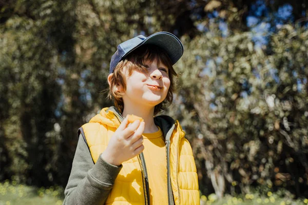 Smiling cheerful child kid in yellow vest and green hoodie eats crisp snacks outdoors in public park. Schoolboy boy enjoying consumes chews junk food outside with trees vegetation on the background — Foto de Stock