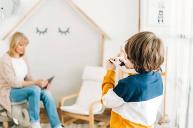 Caucasian school boy kid child taking a photo of his mother mom while she surfing the internet with a tablet. Preschooler playing with camera