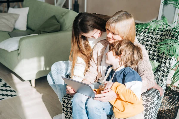Yetişkin kızı ve çocuk çocuğu olan yetişkin bir anne dijital tablet bilgisayar cihazı kullanıyor. Kafkasyalı büyükanne, kız ve torun evde video konferans görüşmesi yapıyorlar.