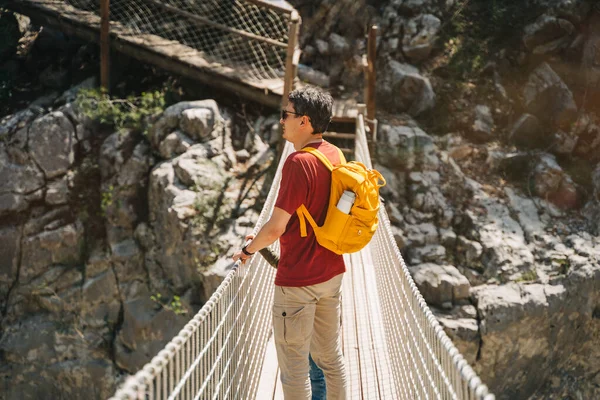 Vater und Sohn schauen beim Wandern von der Hängebrücke in die Berge. Lässig gekleidete Touristen, Schulkind und sein Vater mit gelbem Rucksack überqueren die Schlucht durch eine Hängebrücke — Stockfoto
