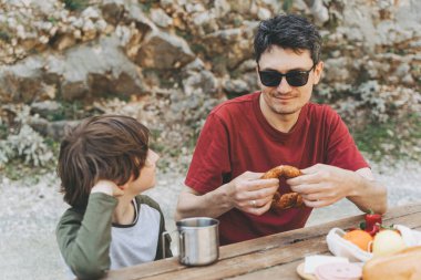 Baba, aile pikniğinde okul çocuğuna sarılıyor. Çocuk çocuk ve babası yürüyüş yaparken dinleniyor ve pikniğin tadını çıkarıyorlar. Çocuk gülüyor ve piknikte ekmeği ısırıyor.