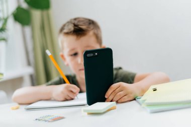 Okula dönelim. Uzaktan öğrenme çevrimiçi eğitim. Kafkasyalı gülümseyen çocuk evde cep telefonuyla ders çalışıyor ve okul ödevi yapıyor. Cep telefonu, kalem ve eğitim kitapları olan bir çocuk.