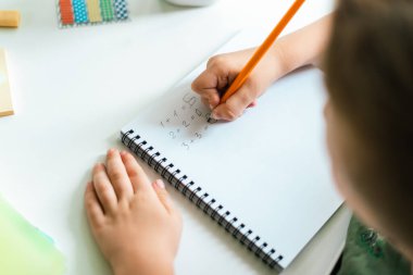 Uzaktan öğrenme çevrimiçi eğitim. Çocuk evde kitapla çalışıyor, not defterine yazıyor ve okul ödevi yapıyor. Çocuk elinde not defteriyle masada oturuyor. Matematik problemlerini çöz. Okula geri dön..