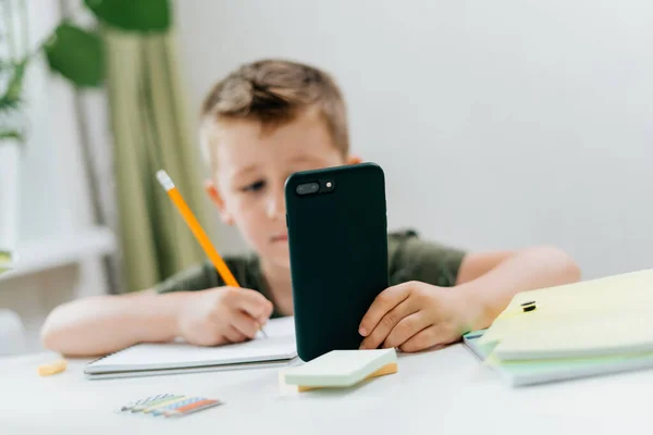Back to school. Distance learning online education. Caucasian smile kid boy studying at home with mobile smart phone and doing school homework. Thinking child with cellphone, pencil and training books