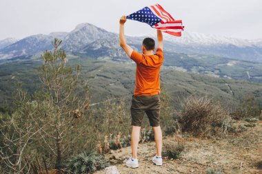 Kayalıklarda duran ve arka plandaki dağlara bakarken Amerikan bayrağını sallayan genç adam. Erkek yolcu dağın tepesinde Amerikan bayrağı sallıyor. 4 Temmuz Bağımsızlık Günü.