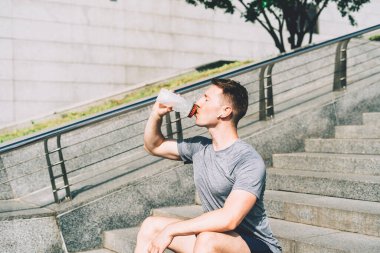 Merdivenlerde oturup spor antrenmanlarından sonra dinlenmekten yorgun düşmüş genç adam. Summer City caddesinde egzersiz yaparken su şişesi içmek.