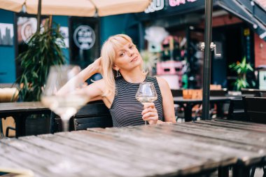 Yetişkin bir kadın dışarıda barda oturmuş şarap bardakları ve bulanık restoran arka planında beyaz şarap içip peynir yiyor. Verandada güneşli bir yaz günü. İnsan yaşam tarzı