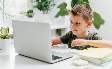 Gülümseyen okul çocuğu evde dizüstü bilgisayarla ders çalışıyor ve ödev yapıyor. Kafkas çocuk elinde defter, kalem ve eğitim kitaplarıyla oturuyor. Okula dönelim. Uzaklık öğrenimi çevrimiçi eğitim. 