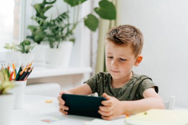 Okullu çocuk evde cep telefonuyla ders çalışıyor ve ödev yapıyor. Okula dönelim. Cep telefonu, kalem ve eğitim kitapları olan beyaz bir çocuk. Uzaklık öğrenimi çevrimiçi eğitim.