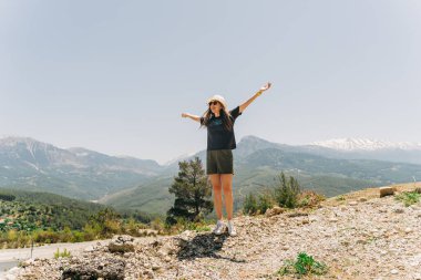 Young woman in hat standing on Mountain View. Raised hands on mountain background.